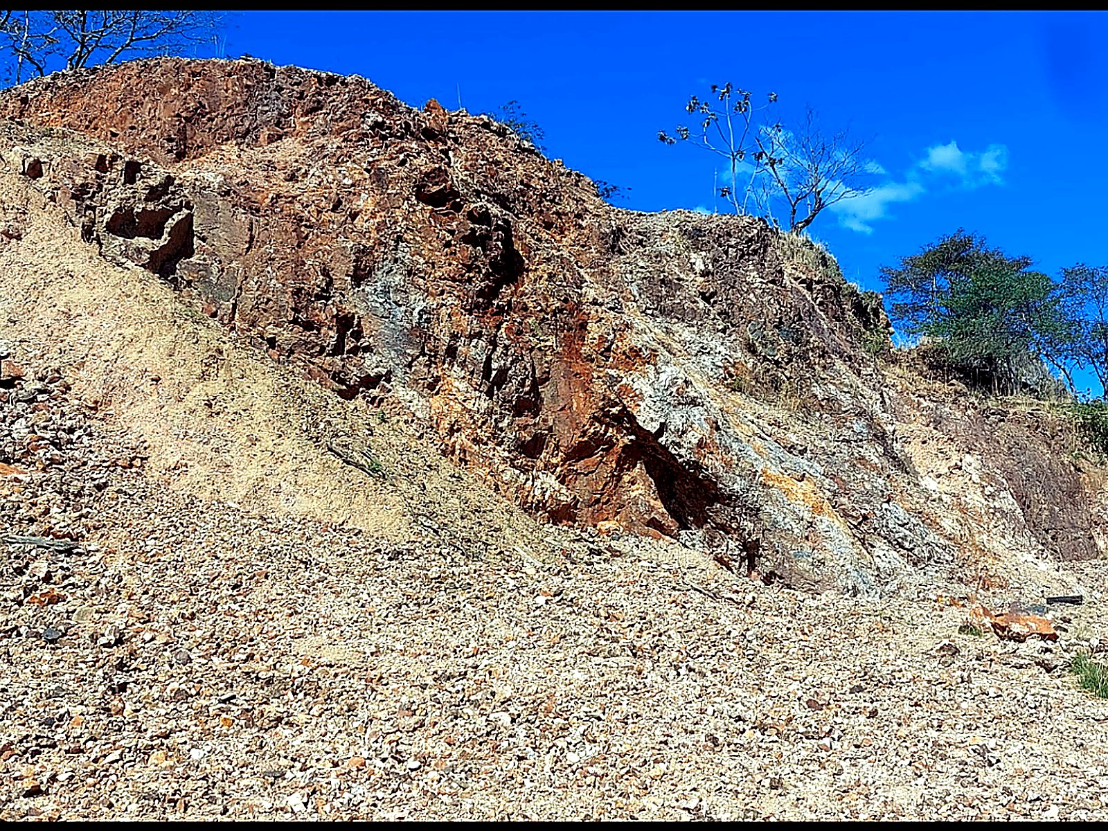 Hillside open cut under blue sky showing a white quartz vein through rust-red oxidised rock