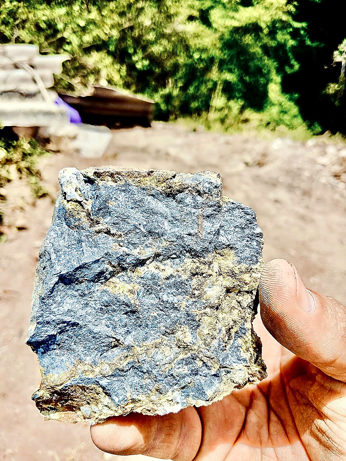 Hand holding a rough rock specimen with a metallic steel-blue antimony face and yellow-brown iron-stained quartz along its edges