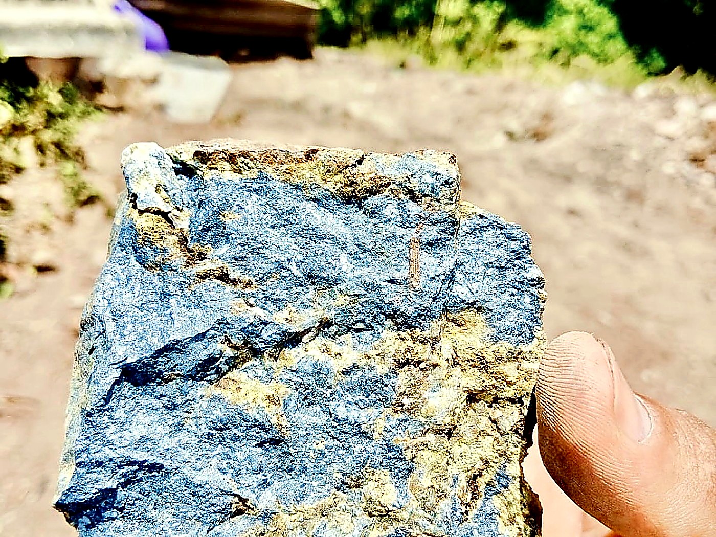 A hand holding a rock sample with a deep blue azurite face and rusty yellow-brown oxidised edges at a mine site