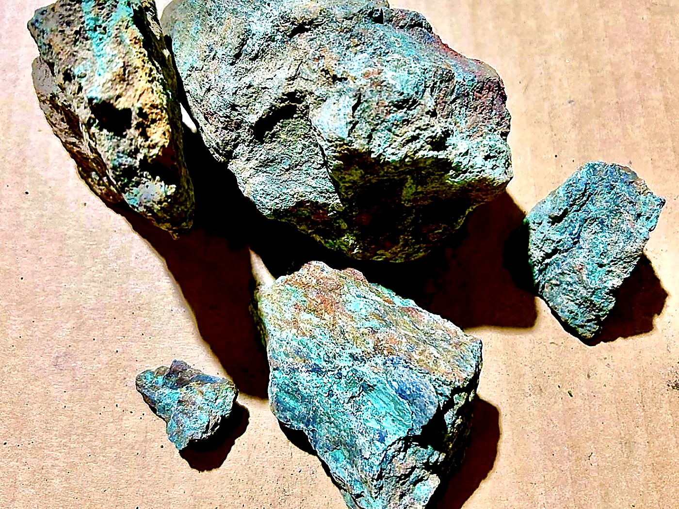Five pieces of rough copper oxide ore with bright turquoise-green malachite and chrysocolla surfaces on a brown paper sheet