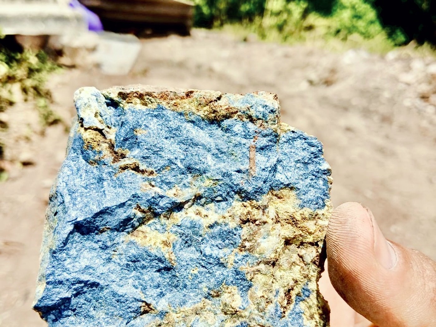 A hand holding a rock sample with a deep blue azurite face and rusty yellow-brown oxidised edges at a mine site
