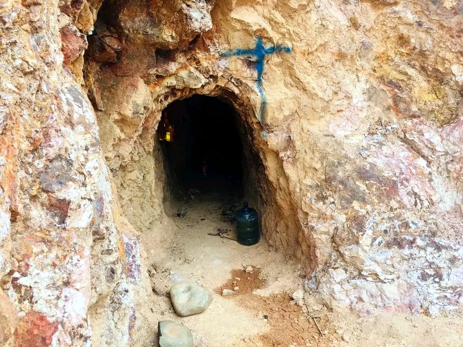 Small arched mine tunnel entrance driven into orange and white oxidised rock, with a blue painted survey cross above the portal