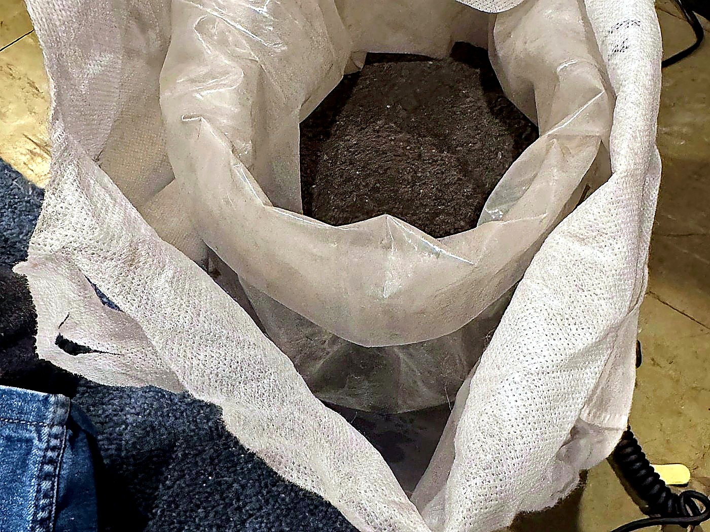 An open white woven sack filled with grey mineral concentrate held open for sampling