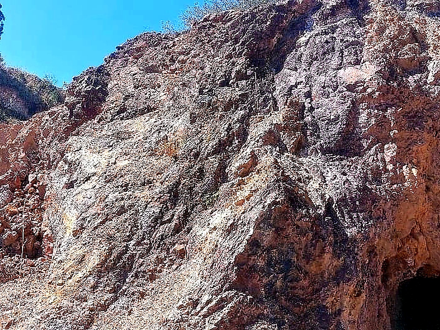 A steep rust-orange and grey oxidised rock face exposing mineralisation against a clear blue sky