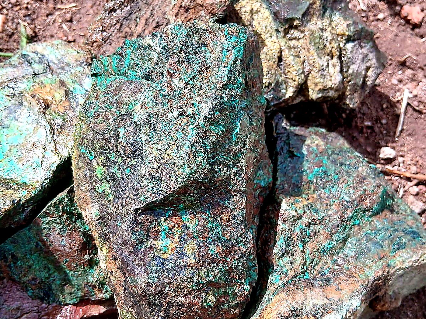 Several chunks of copper ore covered in bright green malachite lying on red-brown soil at a mine site