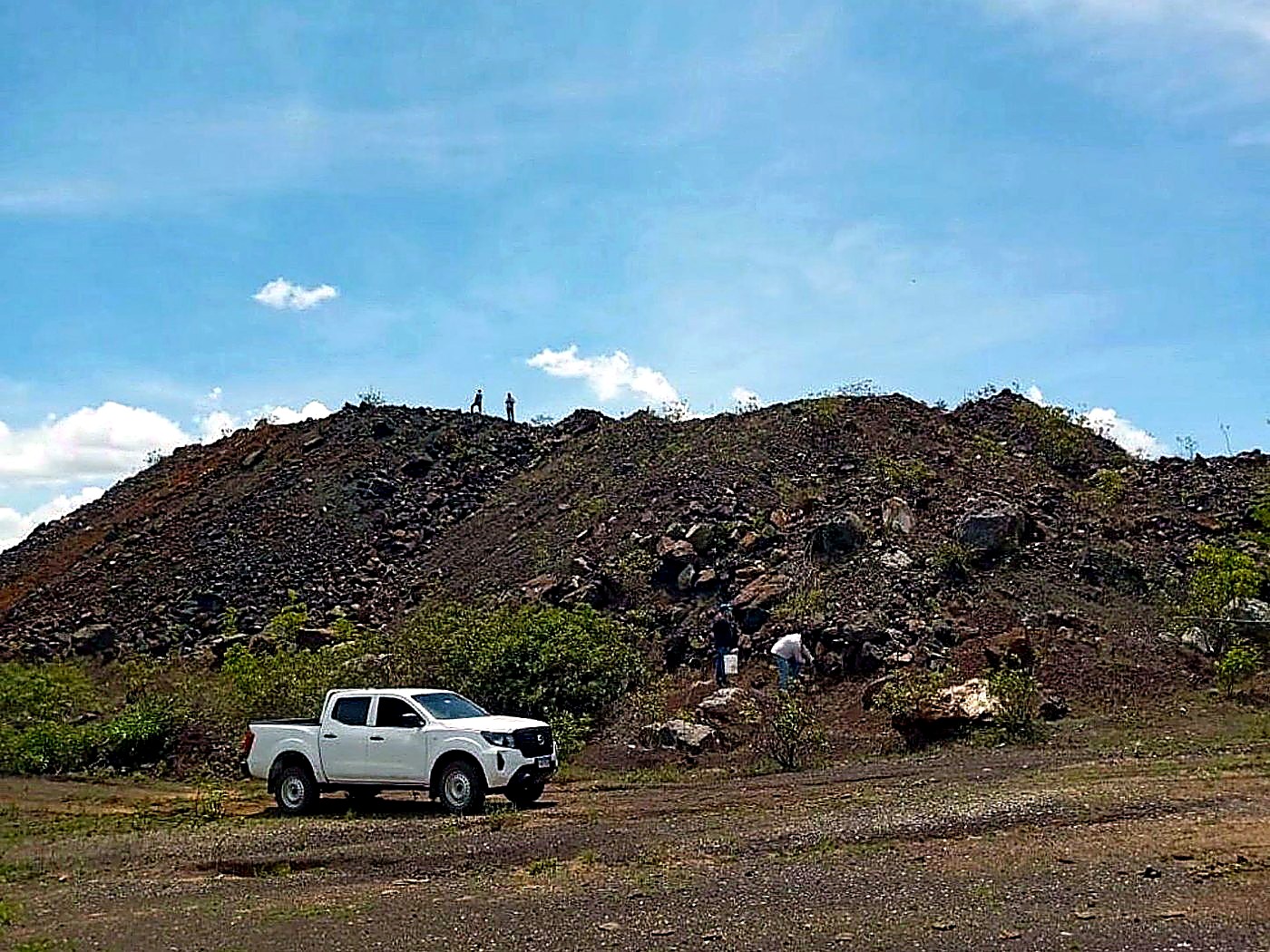 A white pickup truck parked at the base of a dark grey graded ore stockpile on a haulage pad
