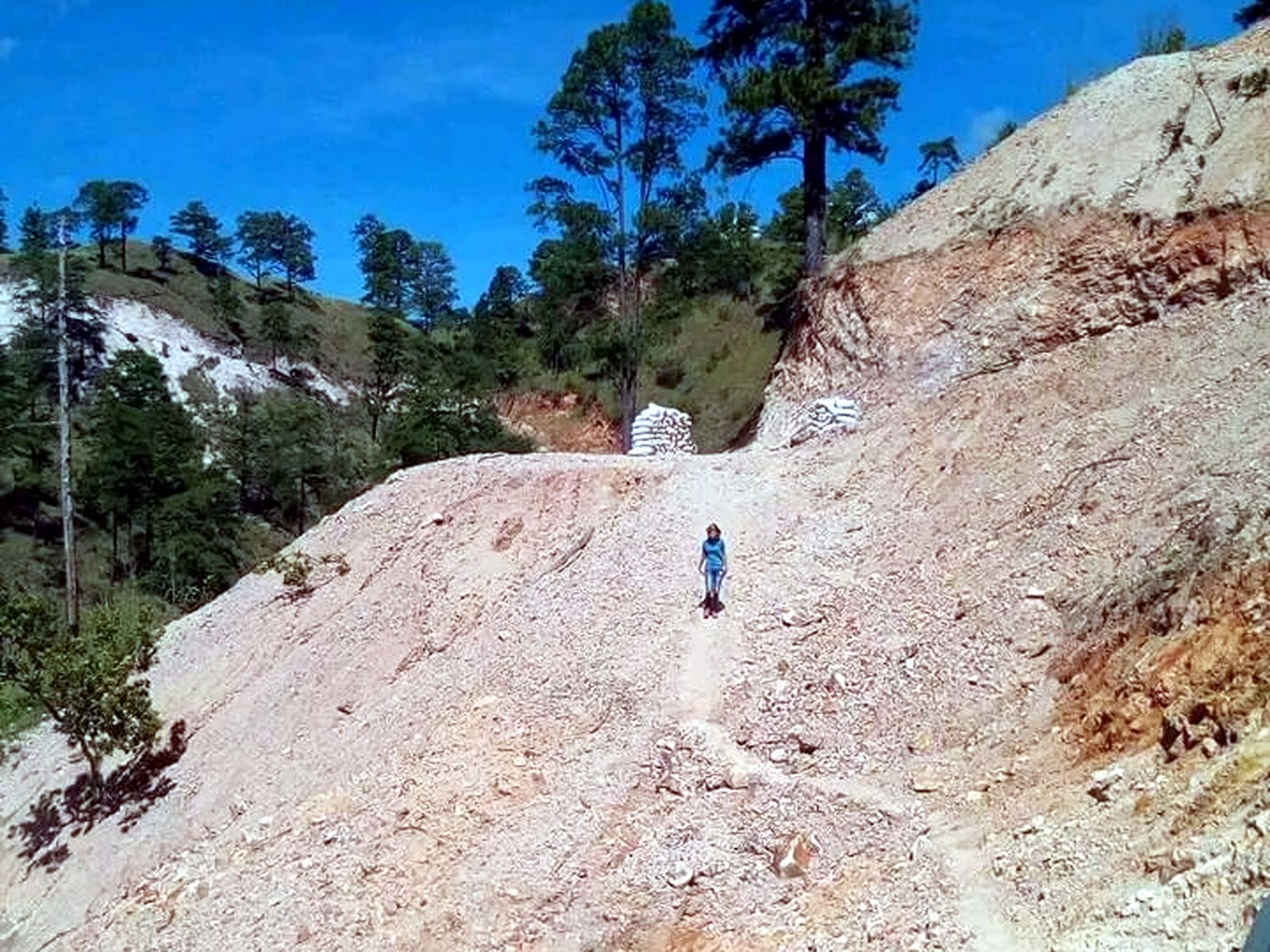 Steep pale rock haul road climbing to a bench where white bulk ore bags are stacked, with a worker walking down