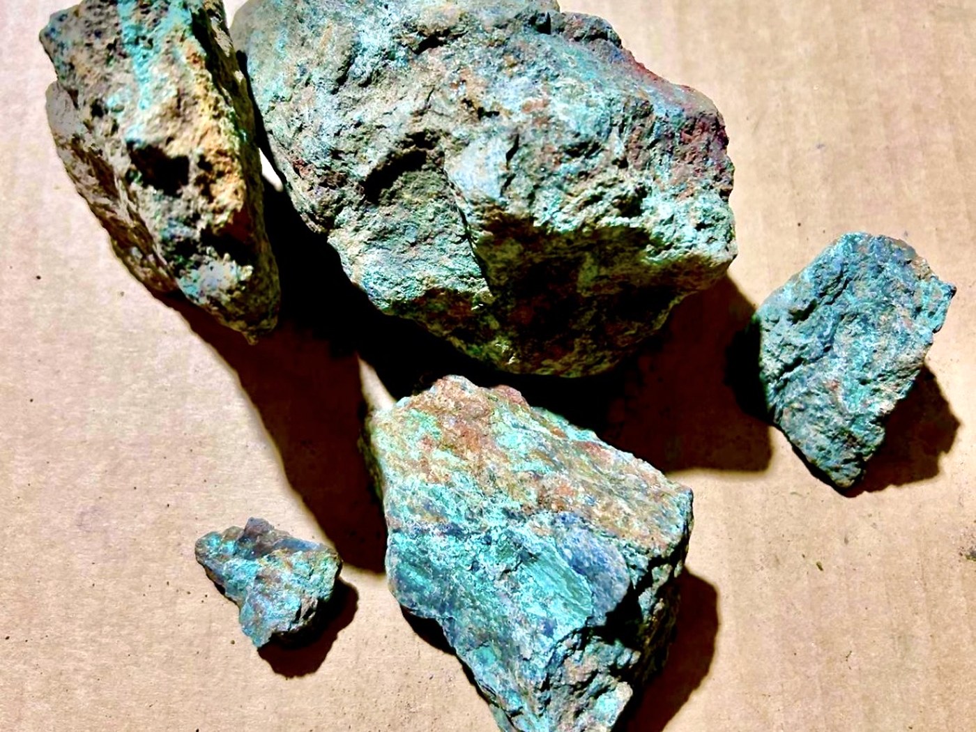 Five pieces of rough copper oxide ore with bright turquoise-green malachite and chrysocolla surfaces on a brown paper sheet