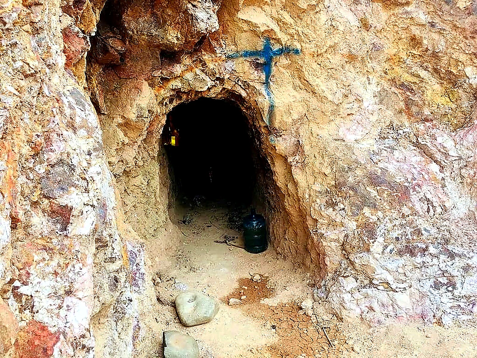 Small arched mine tunnel entrance driven into orange and white oxidised rock, with a blue painted survey cross above the portal